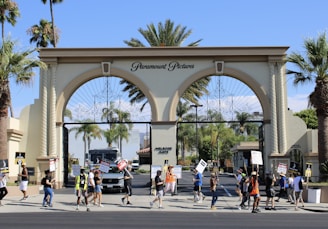 A group of people are holding signs and picketing in front of a large ornate gate with three arches labeled Paramount Pictures. The scene includes palm trees, an entrance sign reading Melrose Gate, and a security vehicle parked inside the gate. The weather appears clear and sunny.