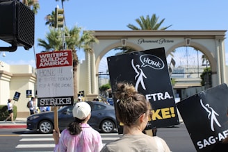 A group of people are participating in a strike near the entrance of Paramount Pictures. They hold signs for the Writers Guild of America and SAG-AFTRA, with messages encouraging cars to honk in support.