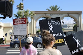 A group of people are participating in a strike near the entrance of Paramount Pictures. They hold signs for the Writers Guild of America and SAG-AFTRA, with messages encouraging cars to honk in support.