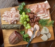 A wooden board is topped with an assortment of deli meats, including slices of prosciutto and stacked piles of various cured meats. A bunch of green grapes is placed alongside sprigs of fresh herbs like parsley and dill. A small knife rests on the board, surrounded by cylindrical slices of pate and several different charcuterie meat styles. Textures and colors give the arrangement a rustic and appetizing presentation.
