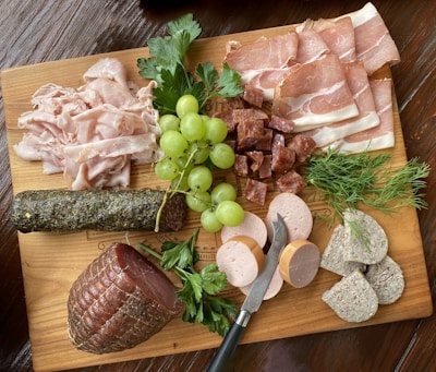 A wooden board is topped with an assortment of deli meats, including slices of prosciutto and stacked piles of various cured meats. A bunch of green grapes is placed alongside sprigs of fresh herbs like parsley and dill. A small knife rests on the board, surrounded by cylindrical slices of pate and several different charcuterie meat styles. Textures and colors give the arrangement a rustic and appetizing presentation.