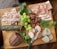 A wooden board is topped with an assortment of deli meats, including slices of prosciutto and stacked piles of various cured meats. A bunch of green grapes is placed alongside sprigs of fresh herbs like parsley and dill. A small knife rests on the board, surrounded by cylindrical slices of pate and several different charcuterie meat styles. Textures and colors give the arrangement a rustic and appetizing presentation.