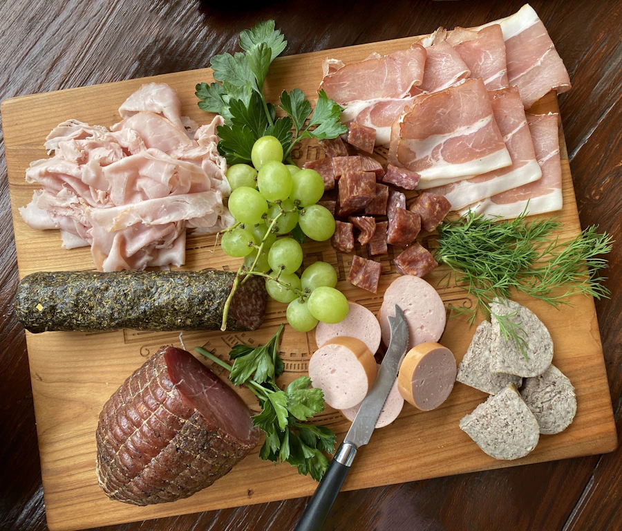 A wooden board of cold cuts, smoked sausage, ham, grapes and dill