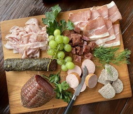 A wooden board is topped with an assortment of deli meats, including slices of prosciutto and stacked piles of various cured meats. A bunch of green grapes is placed alongside sprigs of fresh herbs like parsley and dill. A small knife rests on the board, surrounded by cylindrical slices of pate and several different charcuterie meat styles. Textures and colors give the arrangement a rustic and appetizing presentation.