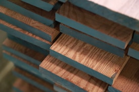 A close-up view of stacked wooden planks with blue-painted edges. The wood grain is visible, and the planks are aligned in a neat, orderly fashion, showing layers of natural wood texture contrasting with painted sections.