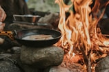 a person cooking food over a skillet over a campfire
