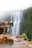 A group of people stands on a viewing platform overlooking a large, cascading waterfall surrounded by lush green vegetation. The scene conveys a sense of natural beauty and grandeur. A small pool of water is visible in the foreground, reflecting some of the surroundings.