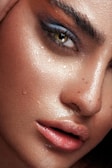 A close-up of a person's face, highlighting a single eye with dramatic makeup. The skin glistens with moisture, showing droplets and a dewy complexion. The makeup consists of bold, dark eyeshadow and thick, defined eyebrows.