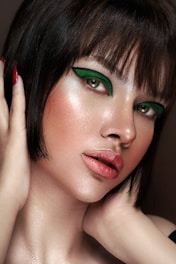 A close-up portrait of a person with short dark hair and striking green eye makeup. The skin has a luminous glow, highlighting the elegant makeup. The lips are painted with a natural shade, and the hands gently touch the face, adding a sense of grace.