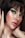 A close-up portrait of a person with short dark hair and striking green eye makeup. The skin has a luminous glow, highlighting the elegant makeup. The lips are painted with a natural shade, and the hands gently touch the face, adding a sense of grace.