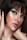 A close-up portrait of a person with short dark hair and striking green eye makeup. The skin has a luminous glow, highlighting the elegant makeup. The lips are painted with a natural shade, and the hands gently touch the face, adding a sense of grace.