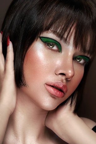 A close-up portrait of a person with short dark hair and striking green eye makeup. The skin has a luminous glow, highlighting the elegant makeup. The lips are painted with a natural shade, and the hands gently touch the face, adding a sense of grace.