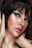 A close-up portrait of a person with short dark hair and striking green eye makeup. The skin has a luminous glow, highlighting the elegant makeup. The lips are painted with a natural shade, and the hands gently touch the face, adding a sense of grace.