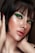 A close-up portrait of a person with short dark hair and striking green eye makeup. The skin has a luminous glow, highlighting the elegant makeup. The lips are painted with a natural shade, and the hands gently touch the face, adding a sense of grace.