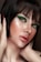 A close-up portrait of a person with short dark hair and striking green eye makeup. The skin has a luminous glow, highlighting the elegant makeup. The lips are painted with a natural shade, and the hands gently touch the face, adding a sense of grace.