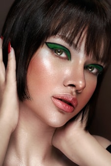 A close-up portrait of a person with short dark hair and striking green eye makeup. The skin has a luminous glow, highlighting the elegant makeup. The lips are painted with a natural shade, and the hands gently touch the face, adding a sense of grace.