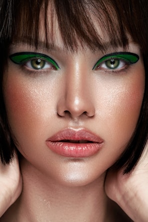 A close-up portrait of a person with striking green eye makeup and well-defined facial features. The skin has a smooth, dewy appearance with natural tones and subtle highlights. The short, dark hair frames the face, accentuating the intense gaze.