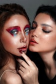 Two individuals are closely positioned, displaying contrasting makeup styles. One has vibrant, artistic makeup with bright red and blue accents around the eyes, and is holding their fingers near their mouth. The other person has a more natural, sleek makeup look with a piercing above their eyebrow.