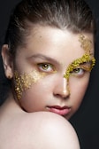 A close-up portrait of a person with artistic gold leaf makeup applied around the right eye and across the cheek. The eyes are intense and striking, accentuated with a hint of yellow. The lighting highlights the skin texture and the gold, creating a dramatic and elegant effect.