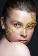A close-up portrait of a person with artistic gold leaf makeup applied around the right eye and across the cheek. The eyes are intense and striking, accentuated with a hint of yellow. The lighting highlights the skin texture and the gold, creating a dramatic and elegant effect.