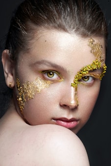 A close-up portrait of a person with artistic gold leaf makeup applied around the right eye and across the cheek. The eyes are intense and striking, accentuated with a hint of yellow. The lighting highlights the skin texture and the gold, creating a dramatic and elegant effect.