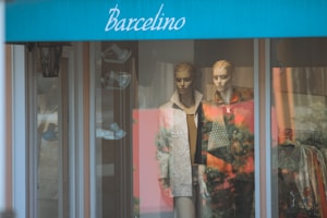 Two mannequins are displayed in a shop window dressed in fashionable clothing, one in a patterned jacket and the other in a textured coat. The store has a blue awning with the word 'Barcelino' on it. Reflections from the glass show outdoor scenery.