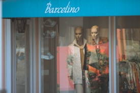 Two mannequins are displayed in a shop window dressed in fashionable clothing, one in a patterned jacket and the other in a textured coat. The store has a blue awning with the word 'Barcelino' on it. Reflections from the glass show outdoor scenery.