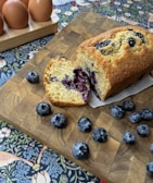 A loaf of blueberry bread sits on a wooden cutting board, with a section cut off to reveal juicy blueberries inside. Fresh blueberries are scattered around the bread. In the background, there is a set of brown eggs in a wooden holder, all placed on a decorative floral-patterned tablecloth.