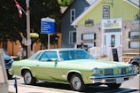 A vintage green car is parked on a street in front of a store with a stone facade and yellow accents. There is a signpost nearby pointing towards the beach, the Bruce Trail, and a campground. The store displays a sign advertising 'apparel, gifts, candies, and toys,' and another shop named 'Dandy Lion' is visible. There are a few people walking in the background and a Canadian flag is attached to a wooden utility pole.