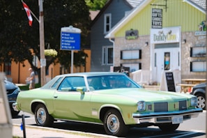 A vintage green car is parked on a street in front of a store with a stone facade and yellow accents. There is a signpost nearby pointing towards the beach, the Bruce Trail, and a campground. The store displays a sign advertising 'apparel, gifts, candies, and toys,' and another shop named 'Dandy Lion' is visible. There are a few people walking in the background and a Canadian flag is attached to a wooden utility pole.