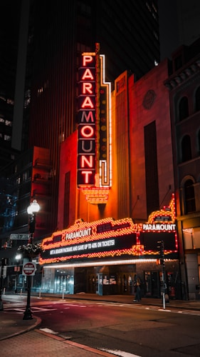 A brightly lit theater marquee with the name 'Paramount' glowing in red and orange neon lights. The entrance is adorned with illuminated signs promoting ticket discounts. The building is set against a backdrop of city structures, and the street in front is quiet and empty at night.