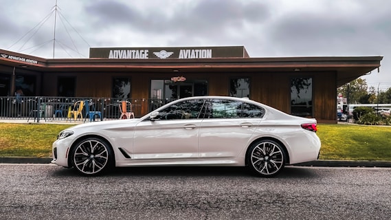 A sleek white car is parked in front of a building with 'Advantage Aviation' written on the facade. The building is wooden with a deck area furnished with colorful chairs. The scene is set under a cloudy sky with visible grass and pavement in the foreground.