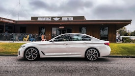 A sleek white car is parked in front of a building with 'Advantage Aviation' written on the facade. The building is wooden with a deck area furnished with colorful chairs. The scene is set under a cloudy sky with visible grass and pavement in the foreground.