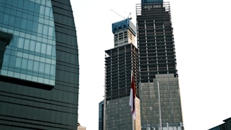 Two skyscrapers under construction stand tall against the sky. One building has a distinctive round shape with glass panels, while the other is a more traditional tall structure with visible cranes at the top. A red and white flag can be seen in the foreground.