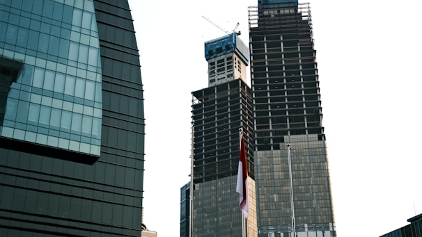 Two skyscrapers under construction stand tall against the sky. One building has a distinctive round shape with glass panels, while the other is a more traditional tall structure with visible cranes at the top. A red and white flag can be seen in the foreground.