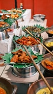 A vibrant display of various dishes served on banana leaves, arranged in metal trays and clay pots. The food includes fried and spicy items, with an assortment of accompaniments around. The setting appears to be a buffet or a festive food setup.