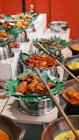 A vibrant display of various dishes served on banana leaves, arranged in metal trays and clay pots. The food includes fried and spicy items, with an assortment of accompaniments around. The setting appears to be a buffet or a festive food setup.