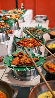 A vibrant display of various dishes served on banana leaves, arranged in metal trays and clay pots. The food includes fried and spicy items, with an assortment of accompaniments around. The setting appears to be a buffet or a festive food setup.