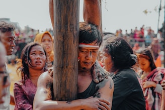 A scene filled with emotional intensity, depicting a group of people engaged in a cultural or ritualistic event. The central figure, with closed eyes and a headband, grips a wooden post with arms smeared in a dark substance. Surrounding them are individuals with expressions of deep emotion, possibly pain or reflection. The background shows an out-of-focus crowd, suggesting a public gathering.