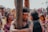A scene filled with emotional intensity, depicting a group of people engaged in a cultural or ritualistic event. The central figure, with closed eyes and a headband, grips a wooden post with arms smeared in a dark substance. Surrounding them are individuals with expressions of deep emotion, possibly pain or reflection. The background shows an out-of-focus crowd, suggesting a public gathering.