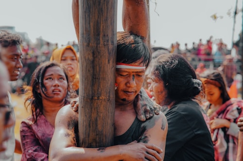 A scene filled with emotional intensity, depicting a group of people engaged in a cultural or ritualistic event. The central figure, with closed eyes and a headband, grips a wooden post with arms smeared in a dark substance. Surrounding them are individuals with expressions of deep emotion, possibly pain or reflection. The background shows an out-of-focus crowd, suggesting a public gathering.