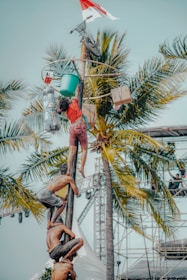 A group of people is climbing a tall, greased pole as part of a competition or festival. They are reaching for items such as a bicycle, boxes, and a bucket at the top of the pole. Palm trees and a stage with scaffolding are visible in the background, indicating an outdoor event. A red and white flag is also seen at the top, likely signifying national or cultural significance.