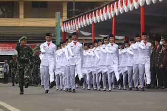 A group of uniformed individuals marches in formation during a parade. They are dressed in white outfits with black hats, and a military figure in camouflage leads them. The event is taking place in an outdoor setting, possibly a ceremonial occasion, with red and white decorations visible.