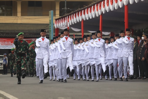 A group of uniformed individuals marches in formation during a parade. They are dressed in white outfits with black hats, and a military figure in camouflage leads them. The event is taking place in an outdoor setting, possibly a ceremonial occasion, with red and white decorations visible.