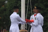 Three individuals in formal white uniforms are engaged in a flag ceremony. They are positioned around a flagpole, holding a folded red flag with precision. Each person wears a black hat, and one of them has a visible emblem on their chest. The background is filled with greenery, suggesting an outdoor setting.