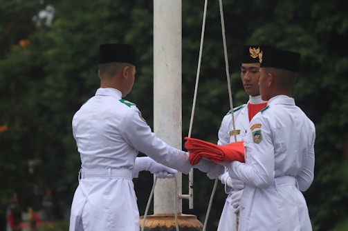 Three individuals in formal white uniforms are engaged in a flag ceremony. They are positioned around a flagpole, holding a folded red flag with precision. Each person wears a black hat, and one of them has a visible emblem on their chest. The background is filled with greenery, suggesting an outdoor setting.