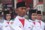 A group of individuals in uniform are participating in a formal parade or ceremony. They are wearing white uniforms with black headgear and have badges on their shoulders, indicating affiliation with an official organization. Their expressions are serious and focused.