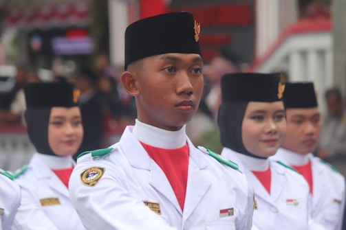 A group of individuals in uniform are participating in a formal parade or ceremony. They are wearing white uniforms with black headgear and have badges on their shoulders, indicating affiliation with an official organization. Their expressions are serious and focused.