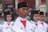 A group of individuals in uniform are participating in a formal parade or ceremony. They are wearing white uniforms with black headgear and have badges on their shoulders, indicating affiliation with an official organization. Their expressions are serious and focused.