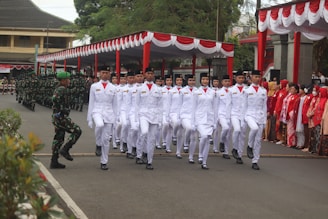 A group of individuals in white ceremonial uniforms are marching in formation. They are part of a formal parade or procession, with soldiers in camouflage uniforms standing in the background. A crowd of people dressed predominantly in red and white traditional attire observes from the side. The setting seems to be outdoors, with a canopy decorated with red and white drapes.